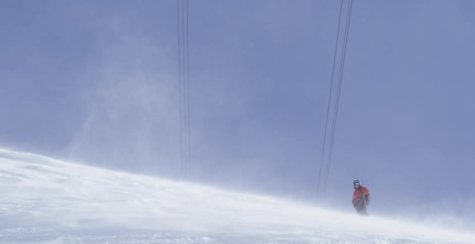 A person stands at the top of a snowy hill as snow blows across