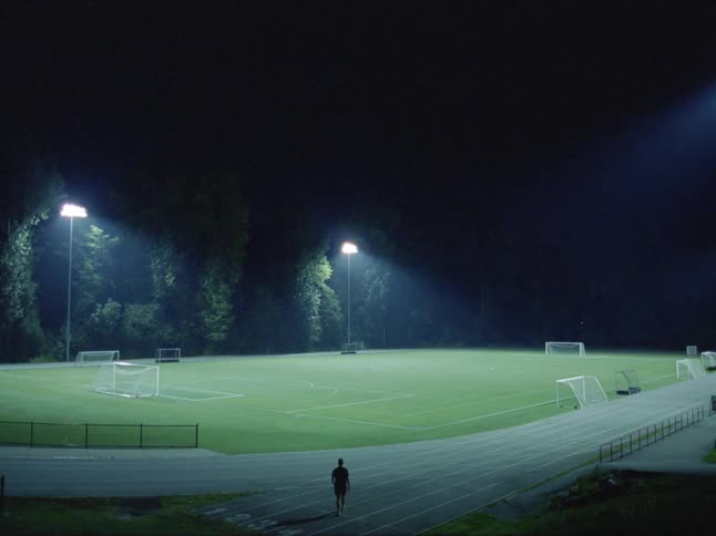 A person walking towards a soccer field with lights and trees in the background