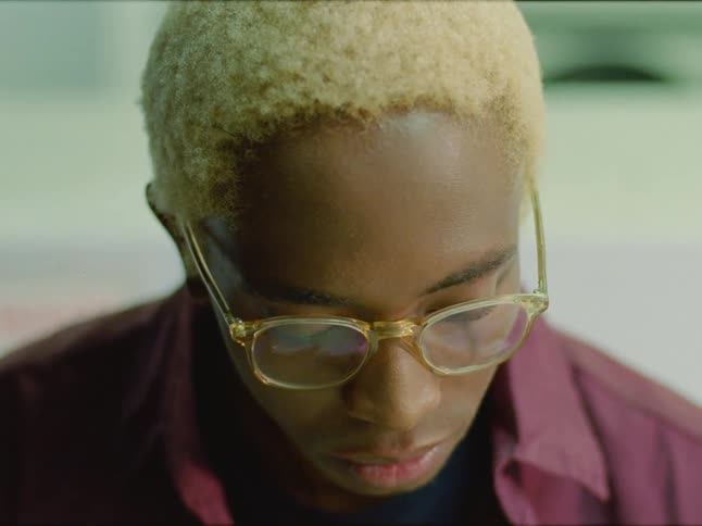 Black man with blonde hair glasses and a red shirt looks down