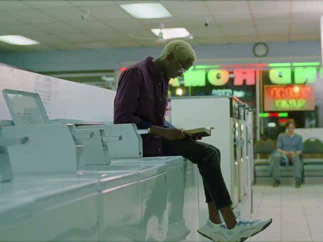 A man with glasses sitting on a washing machine in a laundromat