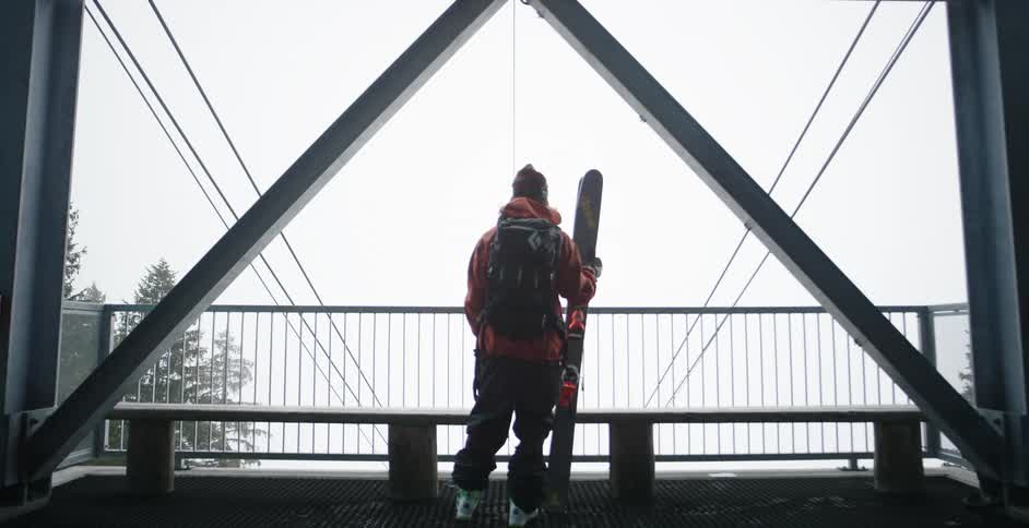 Person stands at top of ski lift structure holding skies and looking out