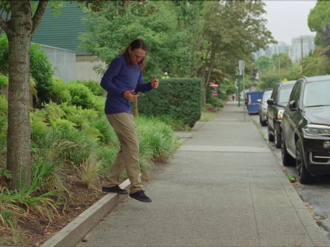 Person hops onto plant bed barrier while looking at mobile phone next to parked cars on street leading from downtown buildings
