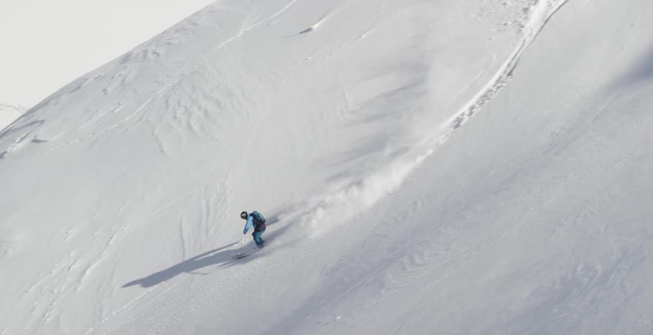 A skier wearing a blue snow suit skis down a steep mountain in a zigzag pattern