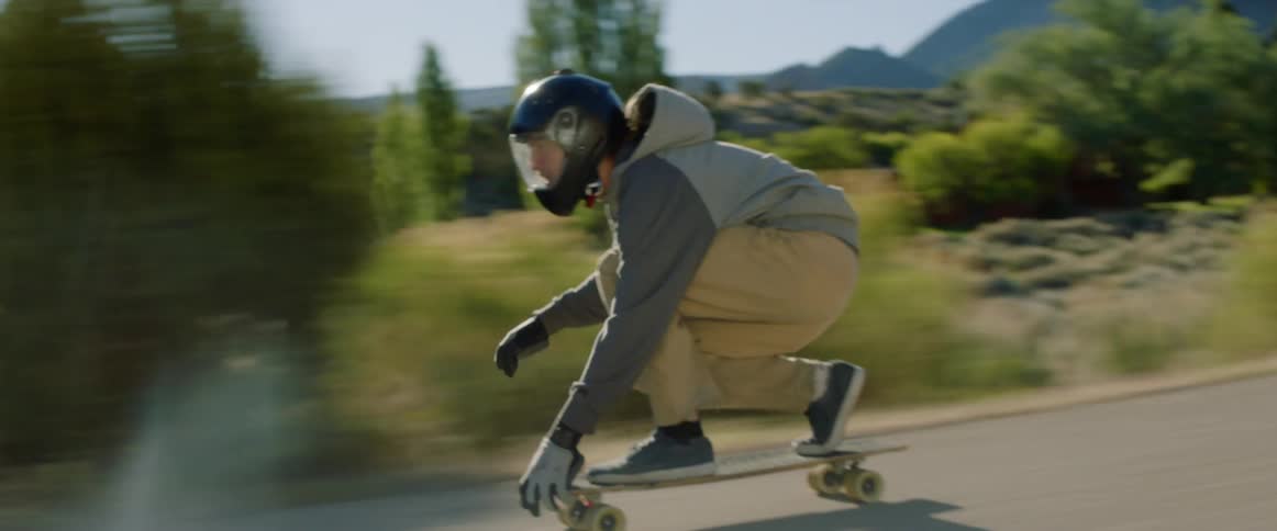 A man rides a skateboard quickly down a sloped road in a rural area near a canyon and hills