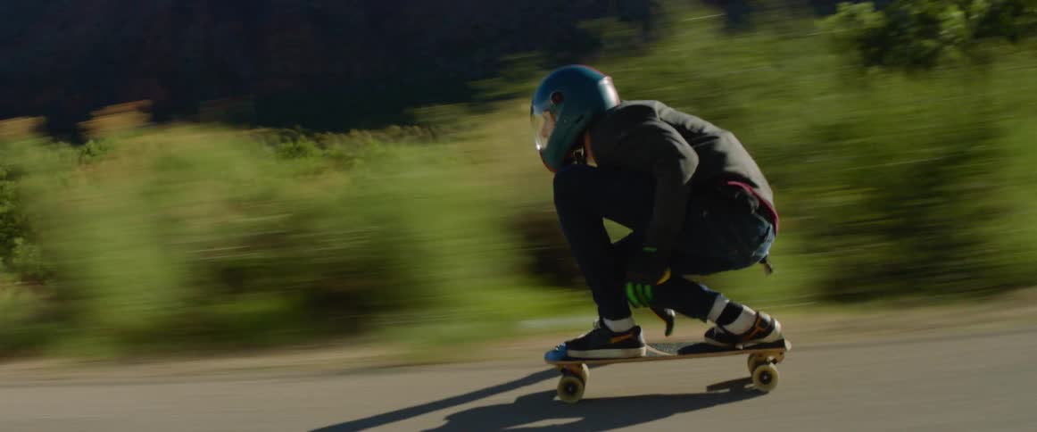 A skateboarder kneels to be aerodynamic as they skate down a countryside road at high speed