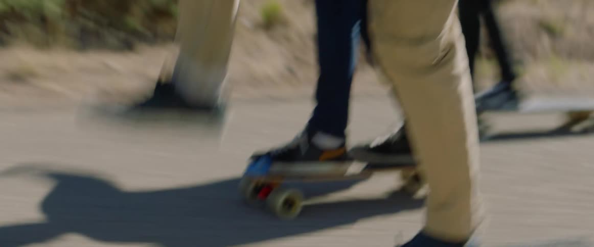 Three people ride skateboards along road past scrub