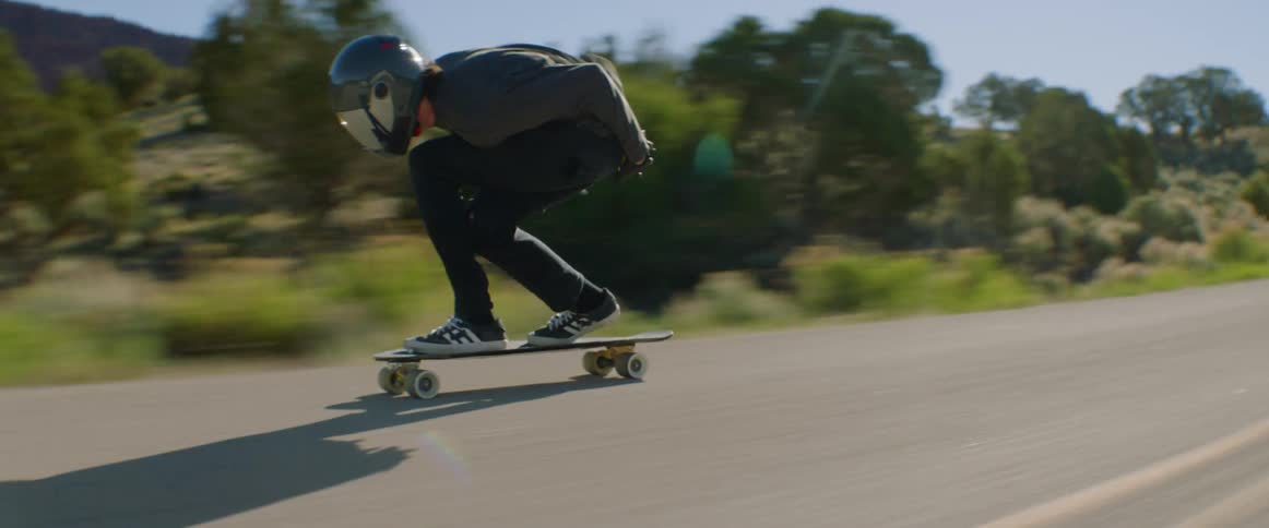 A skateboarder rides down a countryside road at high speed on a sunny day