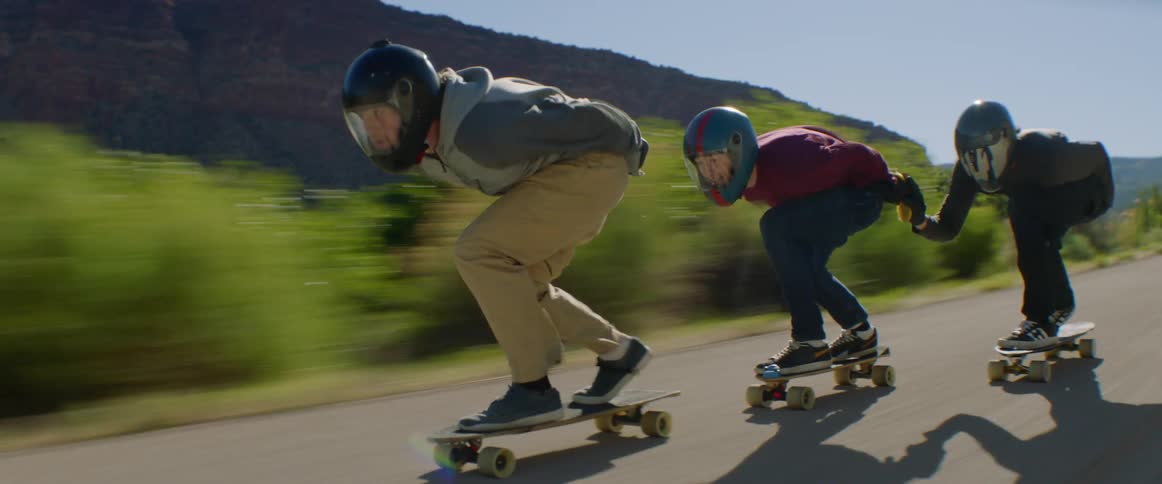Three skateboarders ride along highway past scrub and a mountain