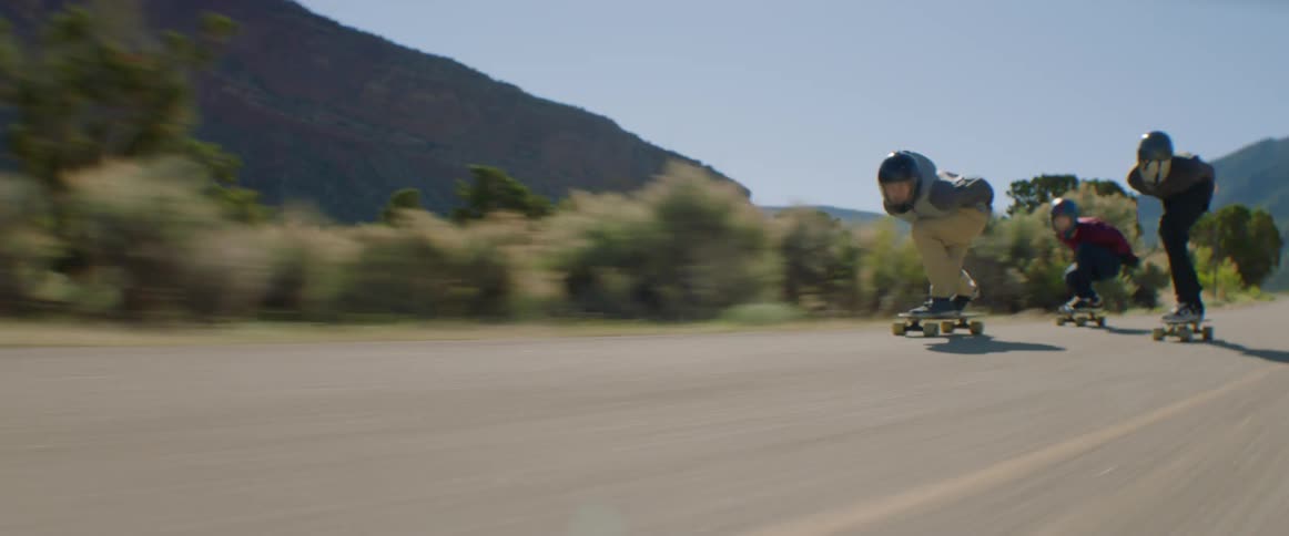 Three people race one another on skateboards down a road