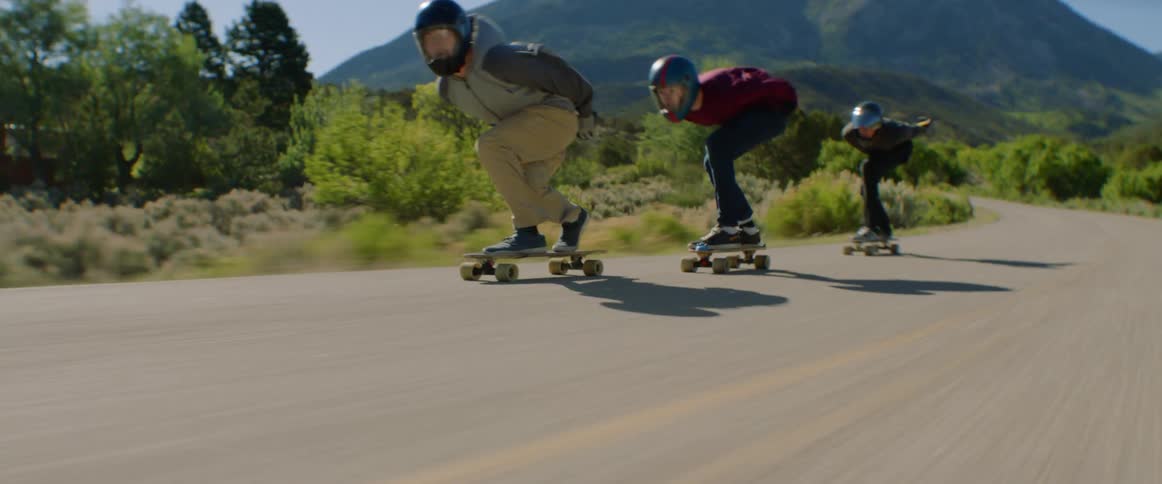 Three men with helmets ride skateboards at high speed on a country road with mountains in the background