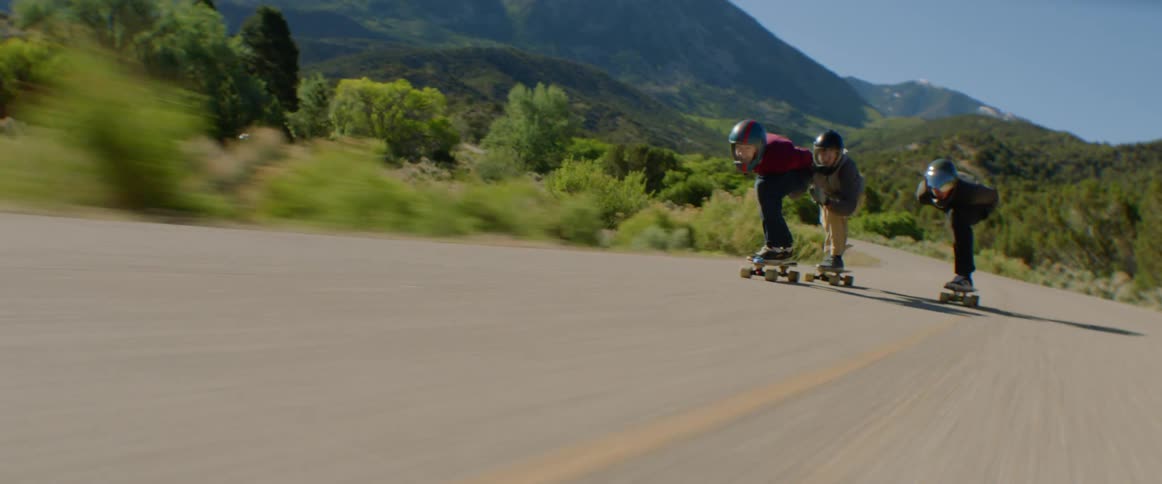 Three teenage skateboarders hold onto each other as they speed down a rural highway