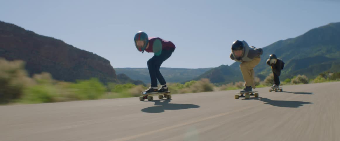 Three skateboarders skateboarding down a road near mountains and hills