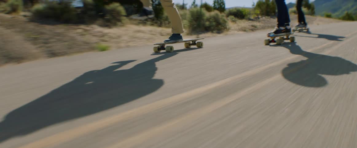 A group of skateboarders skate down a road through the countryside at high speed during the day