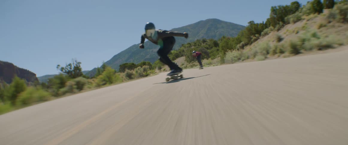 Three skateboarders with helmets ride skateboards at high speed on a country road with mountains in the background