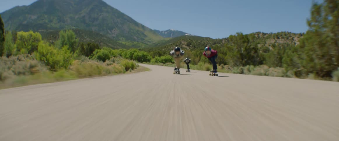 A group of skateboarders race down a road through the countryside at high speed