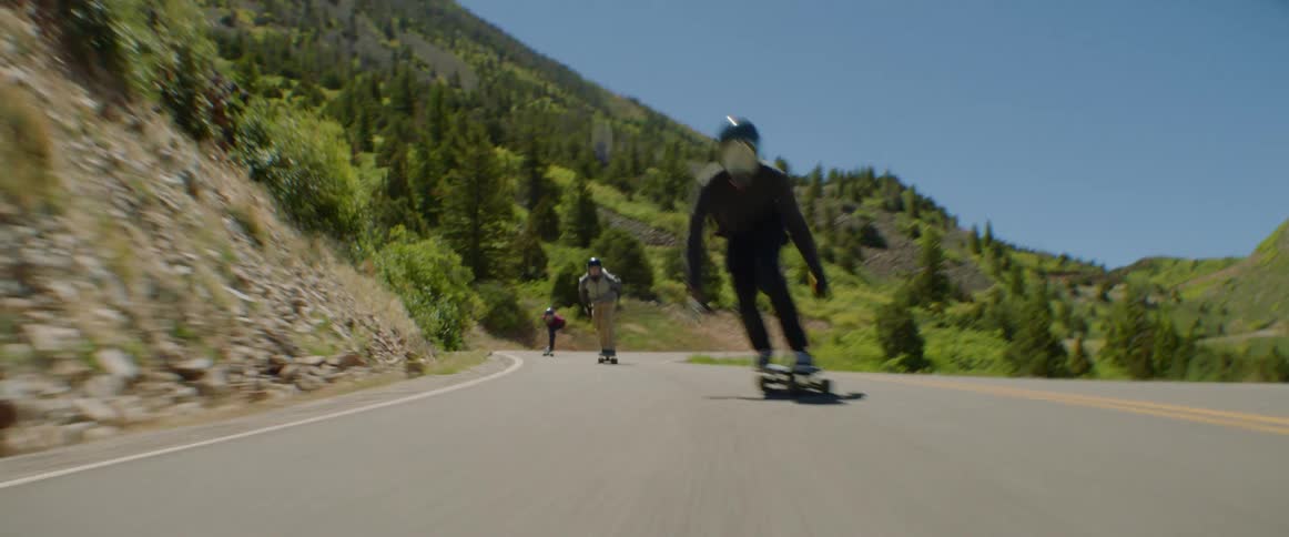 People skateboard along highway past tree covered mountain