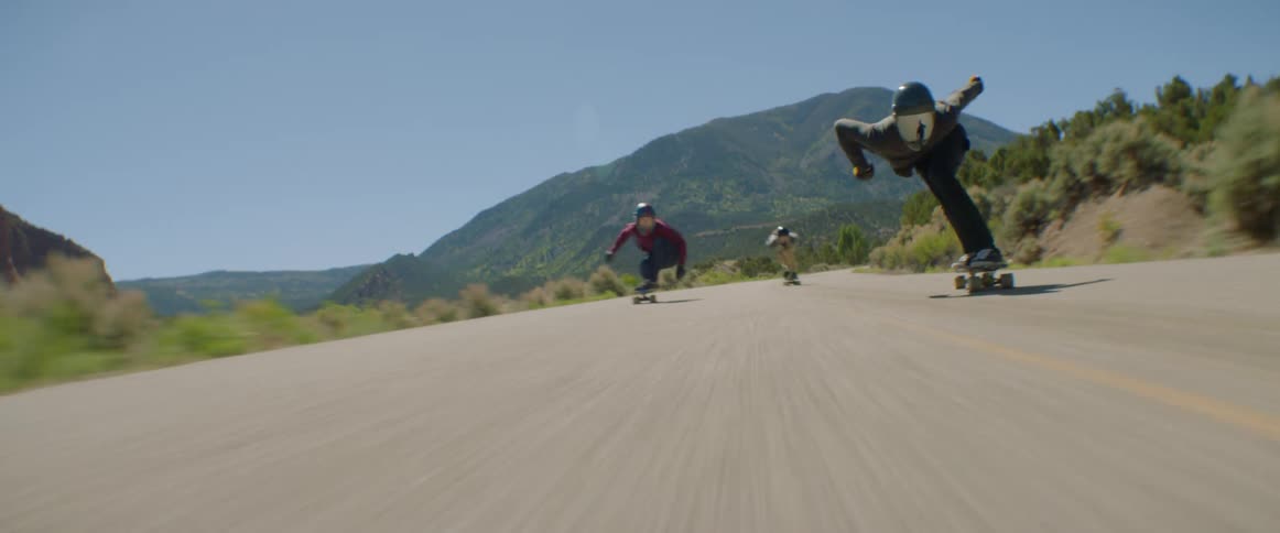 Three skateboarders ride skateboards along highway past scrub and mountains