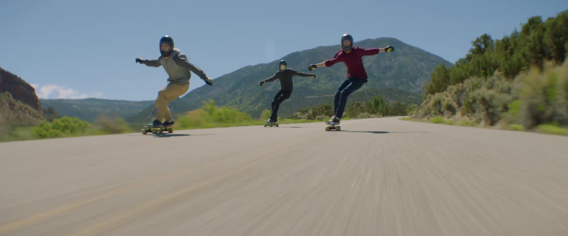 Three men with helmets ride skateboards at high speed on a country road with mountains in the background