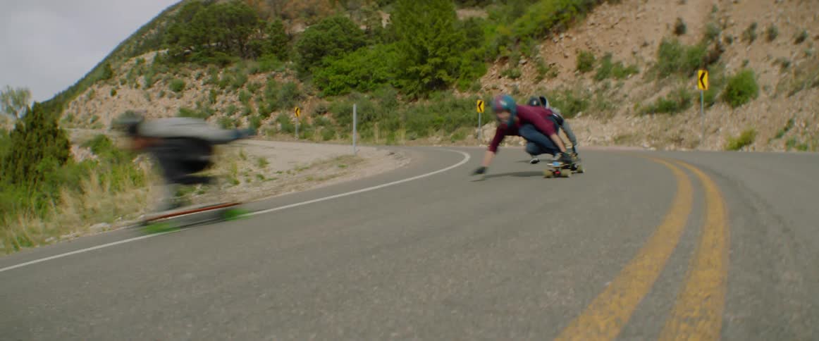 Three people ride skateboards on a rural downhill curved road