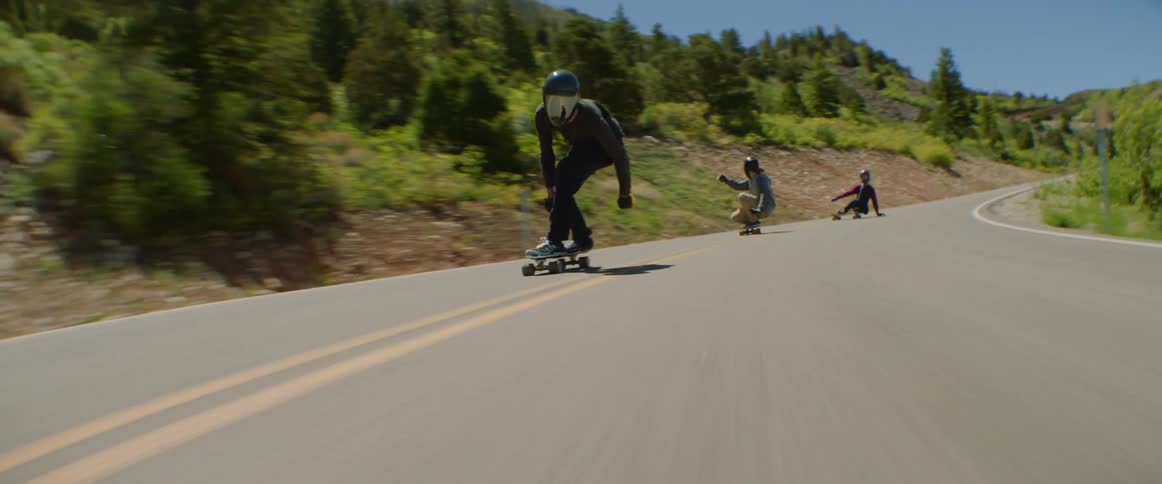 Skateboarders ride skateboards on a winding mountain road with mountains in the background