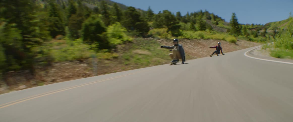 Three skateboarders speeding down a paved road on a mountainside on a summer day