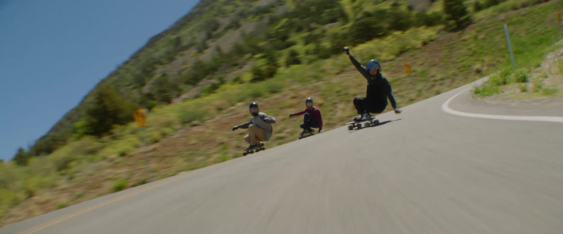 Three people skateboard along highway past scrub and mountains under clear blue sky