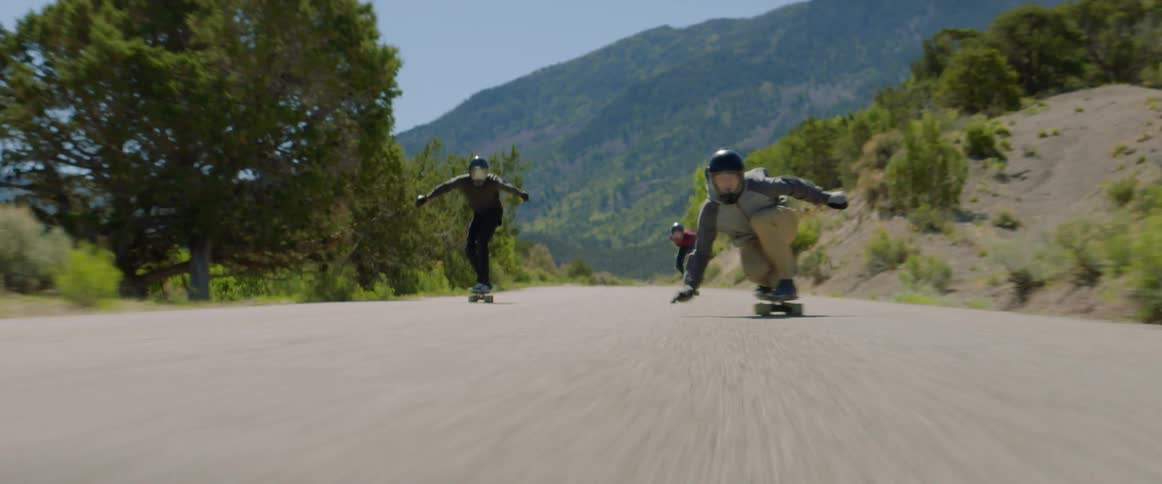 Three people skateboard together on a rural road beside hills and mountains