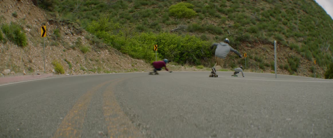 Three skateboarders skateboarding on a road near grassy hills
