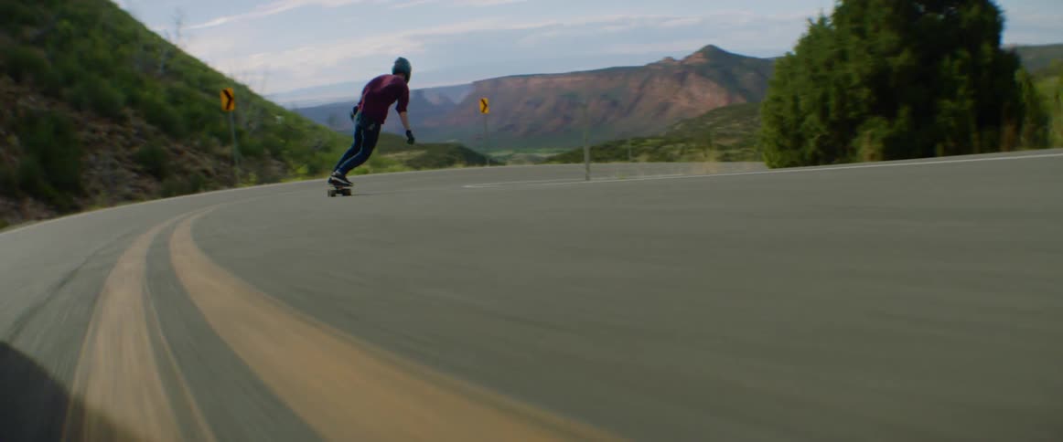 Skateboarder with a helmet rides a skateboard at high speed on a winding road with mountains in the background