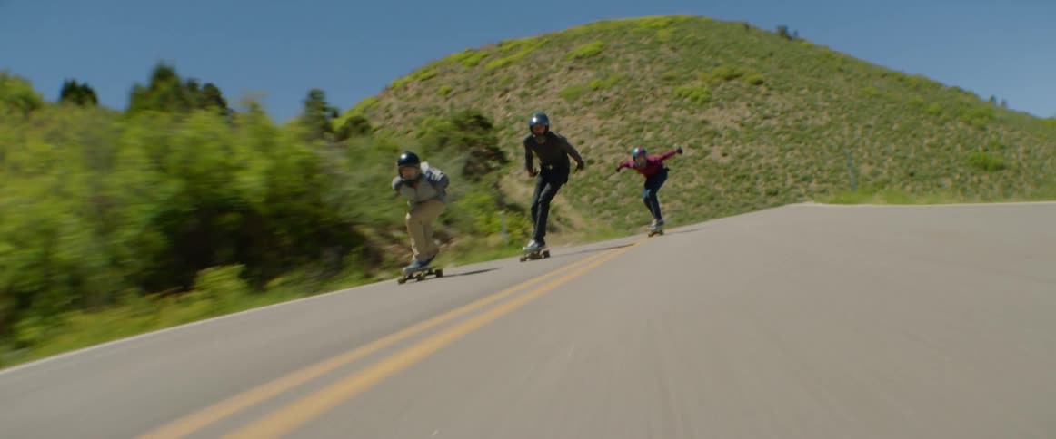A group of skateboarders skate down a road in the countryside at high speed