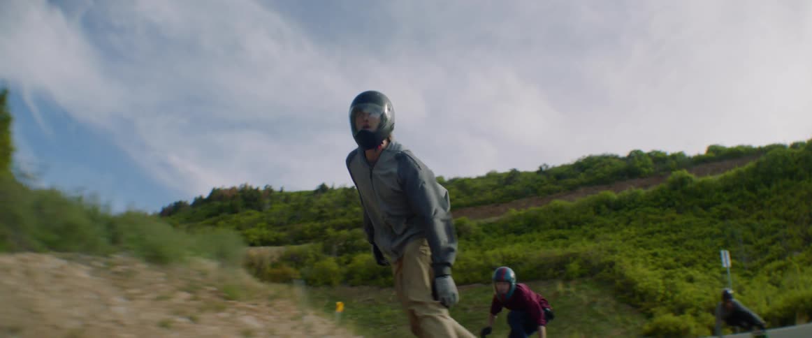 Three people ride skateboards along curving highway past green mountains