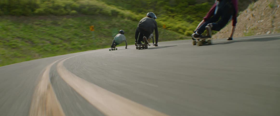 Three teenagers crouch down as they ride skateboards down a curvy highway in a rural area