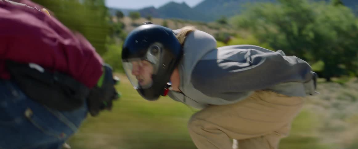 Three people skateboard through countryside past mountains and hills