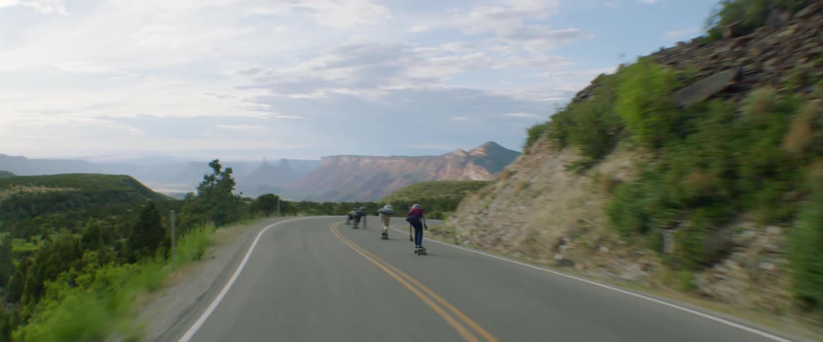 People skateboard along highway past hills and mountains on cloudy day