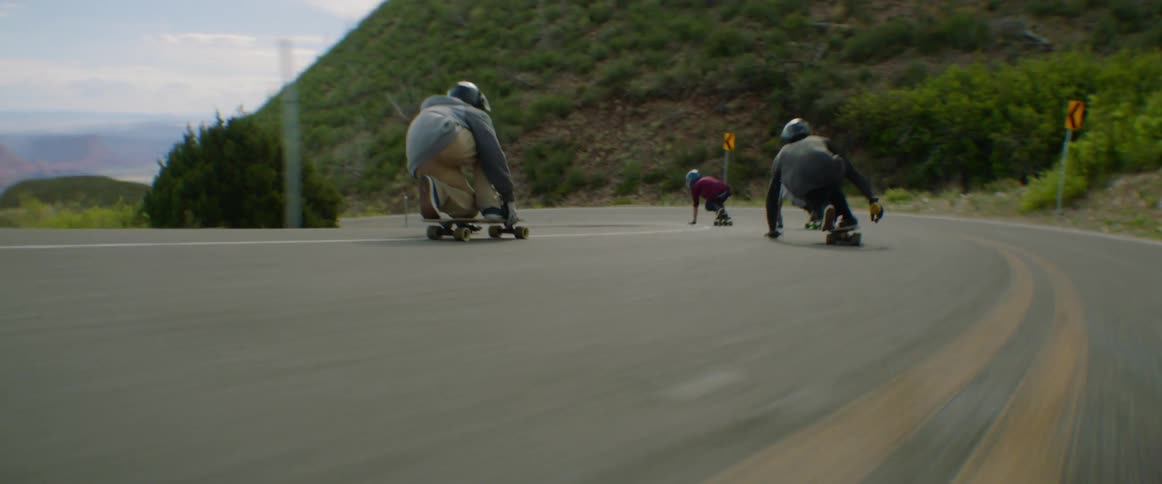 A group skateboarders skate at high speed along a road in the countryside during the day