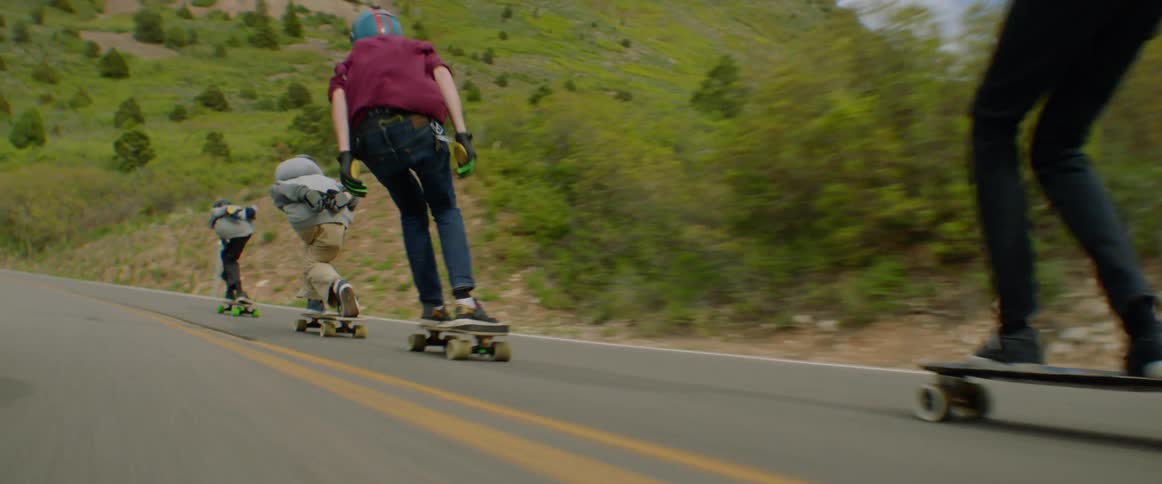 A group of skateboarders skate along a road in the countryside at hill speed