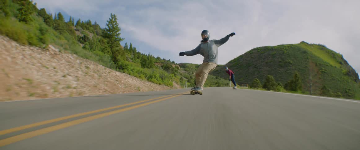 Skateboarders ride their skateboards along highway past mountains slope
