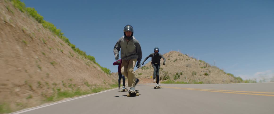 A group of skateboarders kick against the road to gather speed as they descend a road on an incline in the countryside
