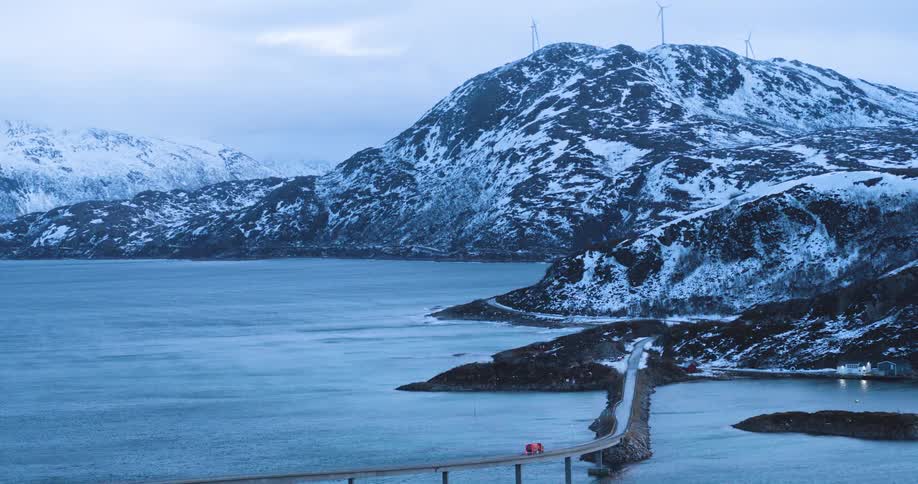 A vehicle driving on a bridge over water with a mountain range the sky and clouds in the background