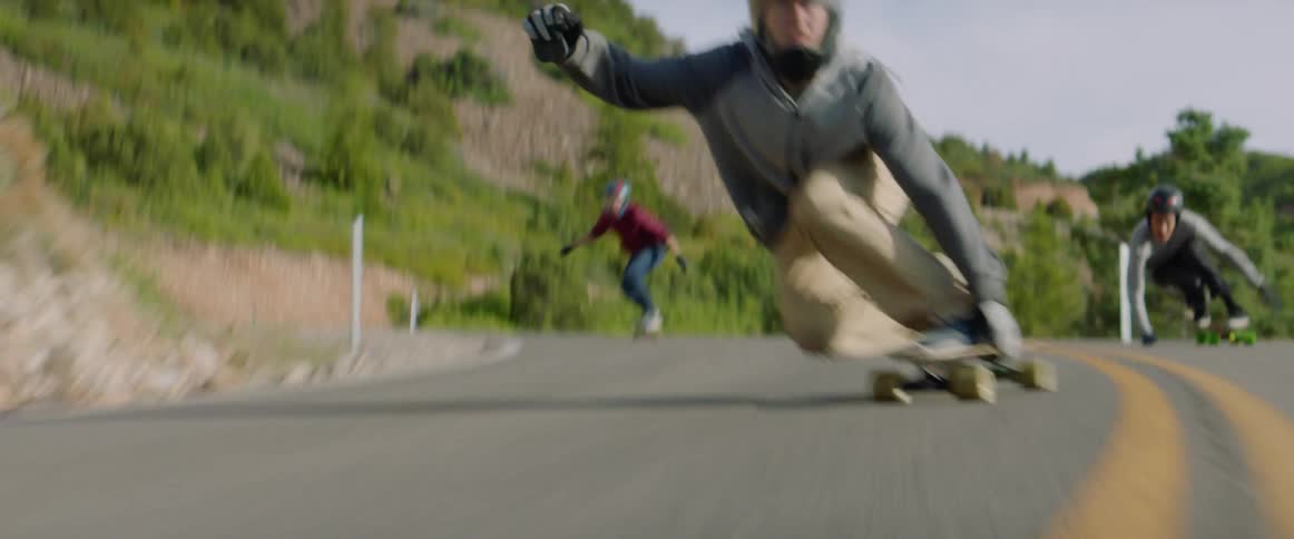 Three people ride skateboards on a quiet rural hillside road