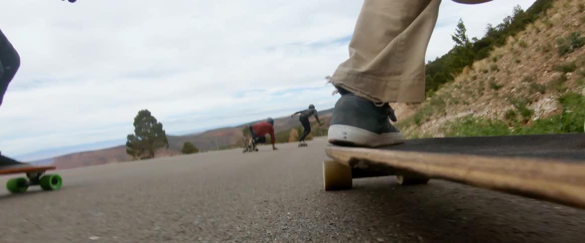 Skateboarders skate along road past countryside and slopes performing tricks