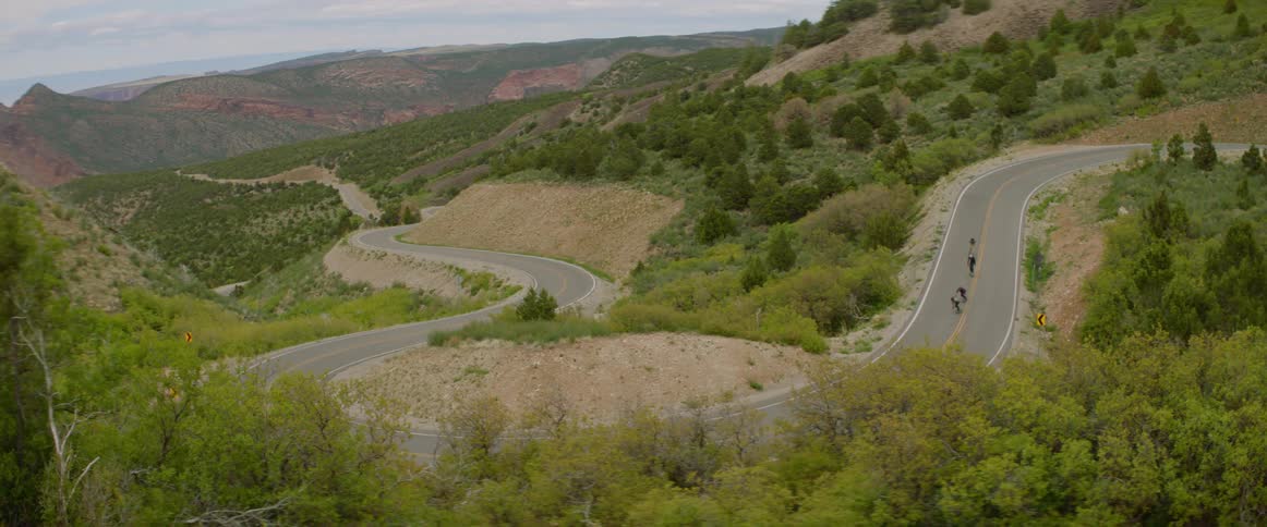 A group of skateboarders travel at high speed around a sharp turn on a road through the hills in the countryside