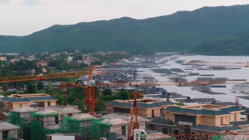 Construction cranes tower over roofs of apartment buildings near lagoon with misty mountains on horizon