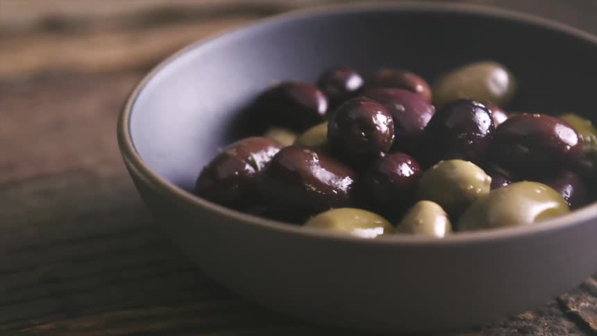 A bowl of olive sits on a wooden table along with bread and cheese