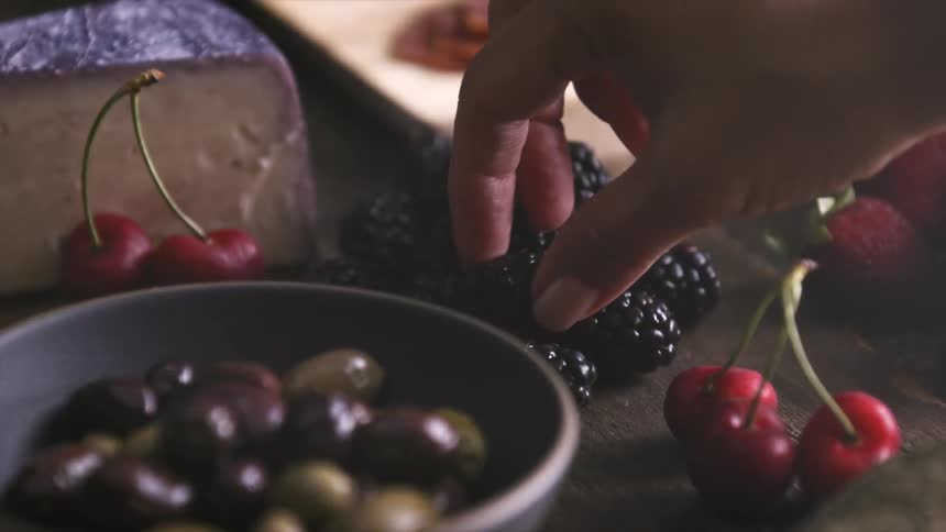 A person places raspberries on a wooden table next to cherries and cheese