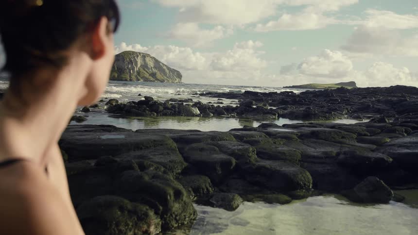 A young woman sits on a rock formation on a beach while looking gazing out over the ocean on a sunny day