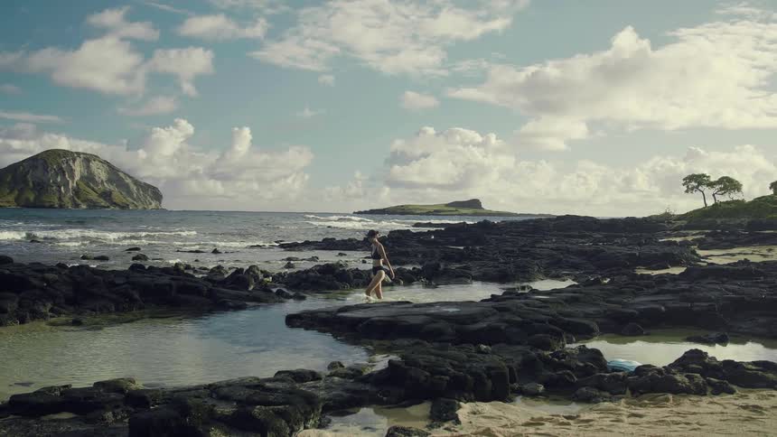 A woman in a bikini wading in water on a rocky beach