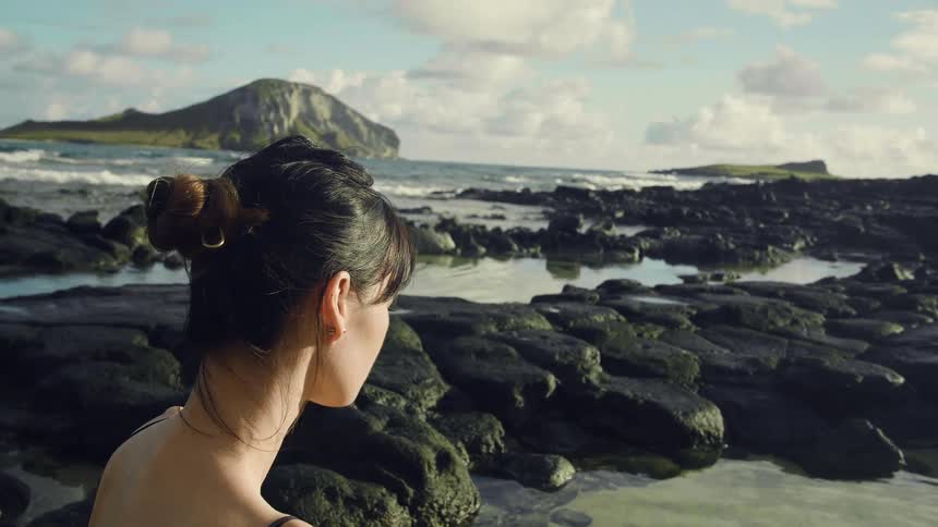 A young woman sits on the rocky coast and gazes out at the incoming tide on a sunny day