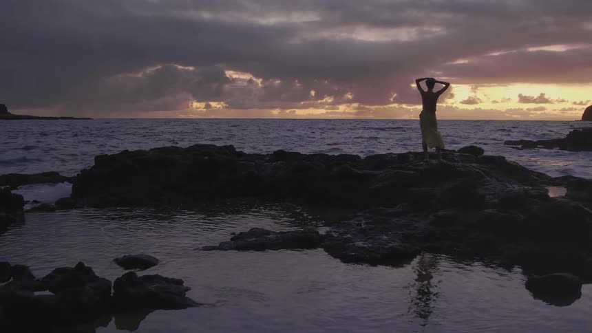 The water from an ocean wave splashes on a woman standing on a rock formation on a beach at sunset