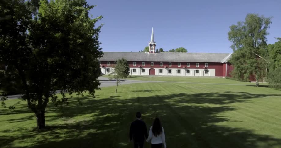 Two people walk hand-in-hand across a grassy lawn towards a building